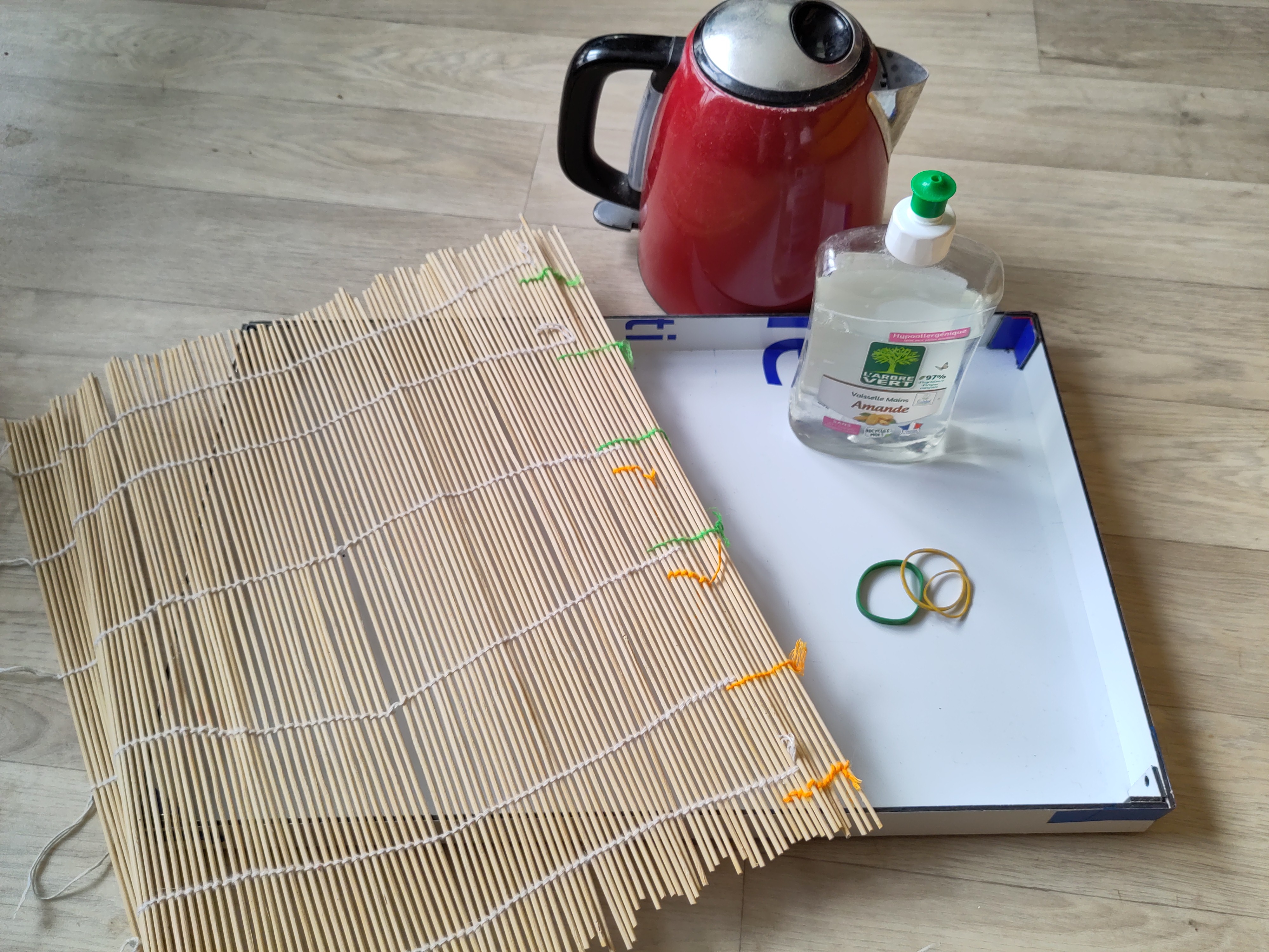 un plateau blanc avec une nate en bambou, des élastiques et du liquide vaisselle dessus. il y a un bouilloire rouge en fond.